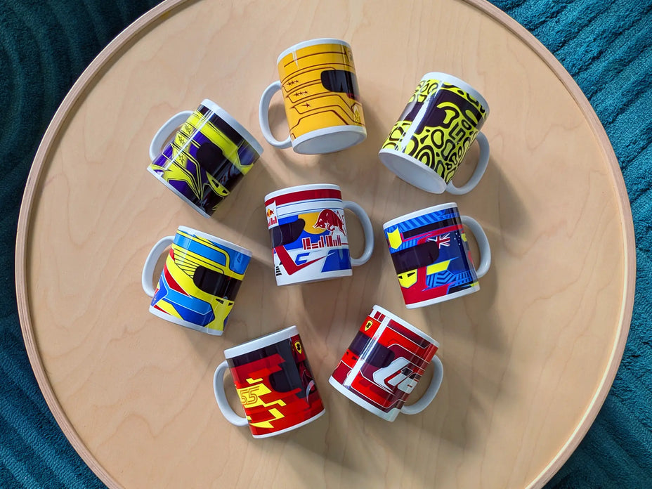 Collection of eight F1-themed mugs featuring minimalist helmet designs inspired by Lewis Hamilton, Max Verstappen, Lando Norris, Oscar Piastri, Fernando Alonso, Carlos Sainz, and Charles Leclerc, displayed on a round wooden table.
