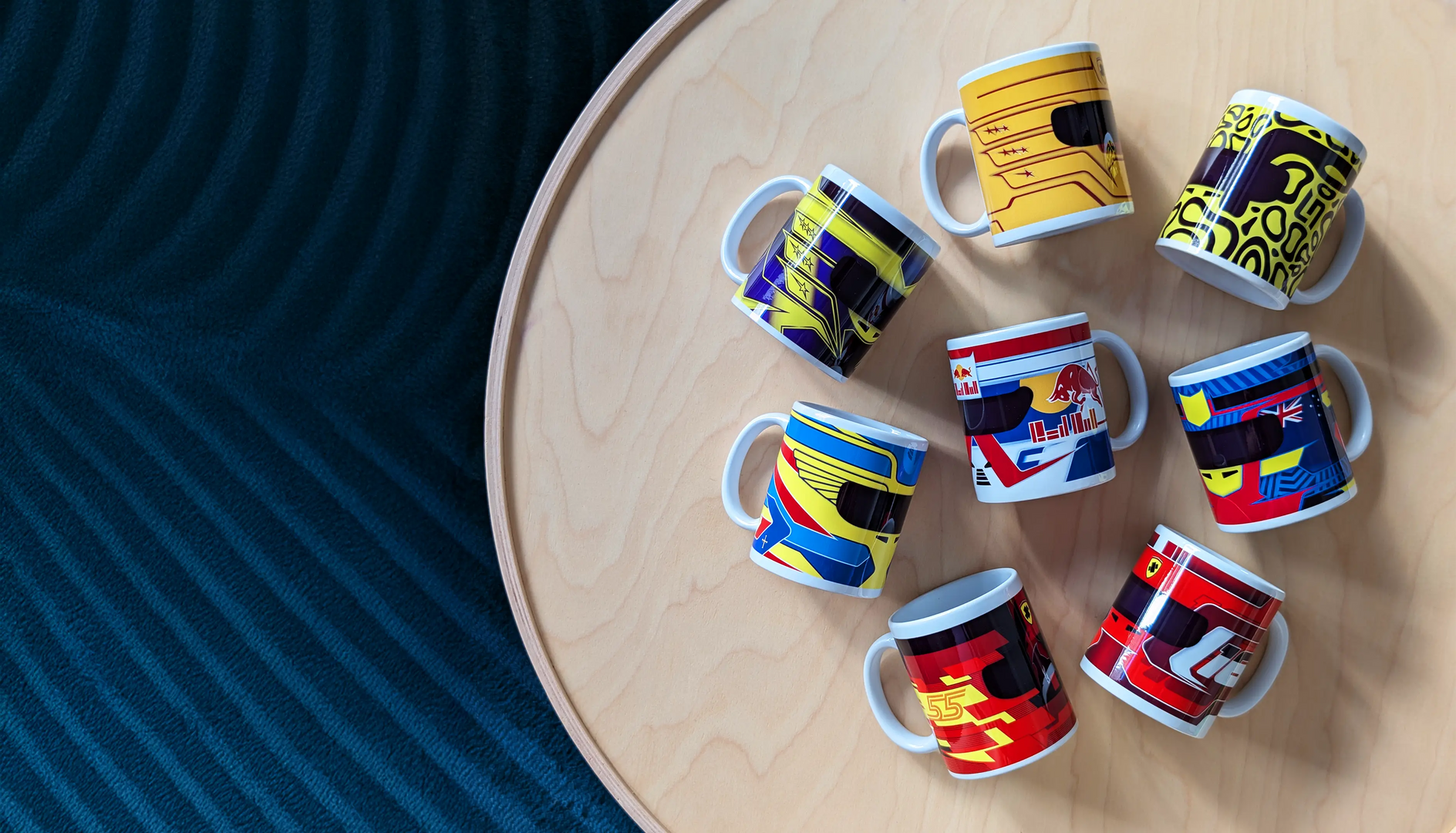 Collection of eight F1-themed mugs featuring minimalist helmet designs inspired by Lewis Hamilton, Max Verstappen, Lando Norris, Oscar Piastri, Fernando Alonso, Carlos Sainz, and Charles Leclerc, displayed on a round wooden table.